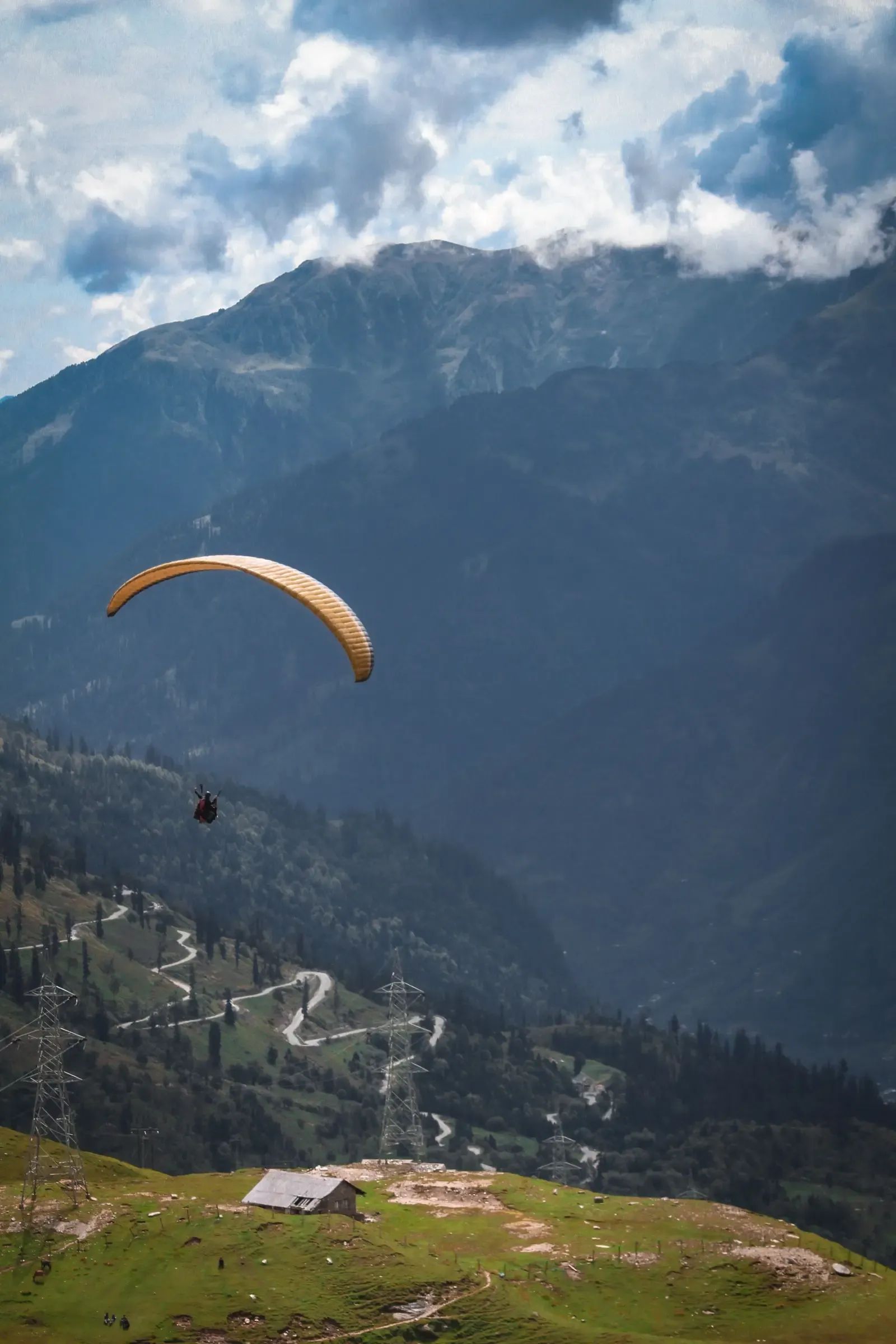 A paraglider turning above the green ridge at Billing, the valley wall rising dark behind and a switchback road far below.