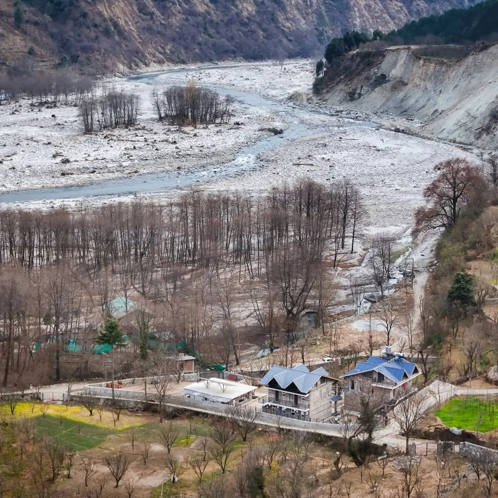 Bare winter trees along a wide stony riverbed with a blue-roofed building on the far bank and snow on the ground.