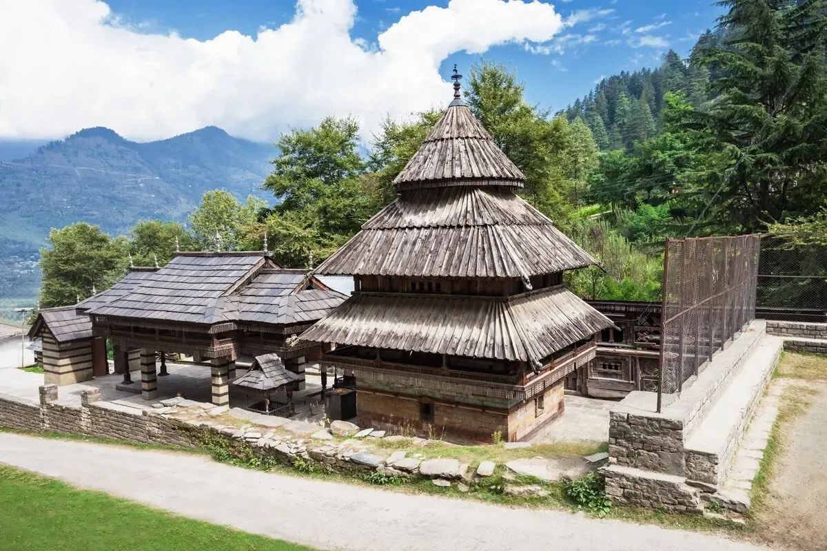 A tiered wooden temple with a conical shingled roof and carved eaves, standing on a walled terrace above the valley in deodar forest.