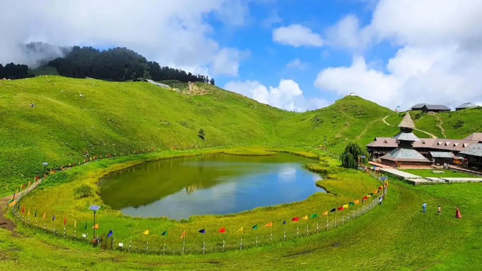A three-tiered pagoda temple standing on a green bowl of meadow beside a small dark lake, forest closing the ridge behind.