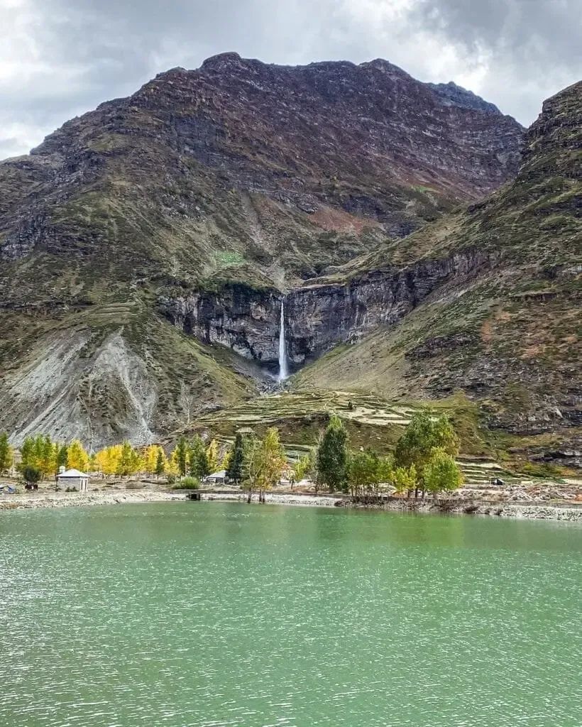 A waterfall dropping the full height of a dark layered cliff, with terraced fields and a line of autumn poplars on the slope below it.